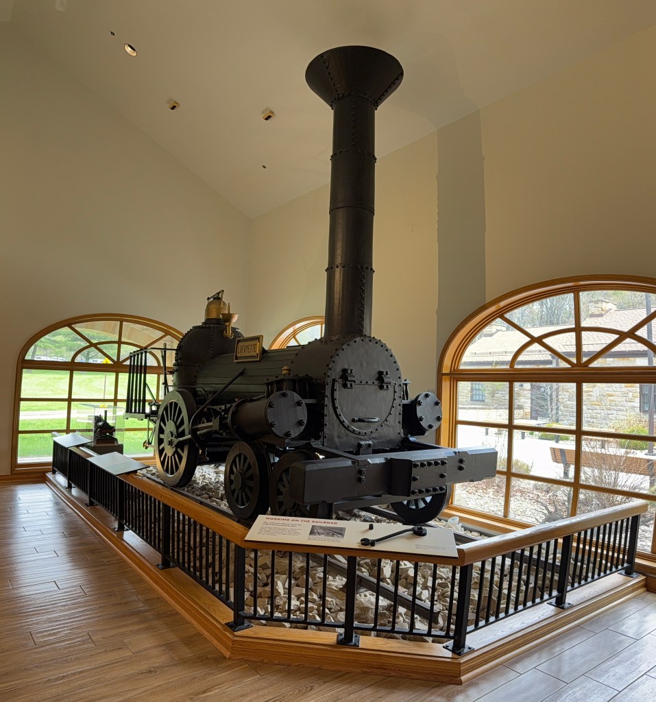 A historic steam locomotive displayed indoors, surrounded by wooden railings and large windows showcasing the outdoor scenery.