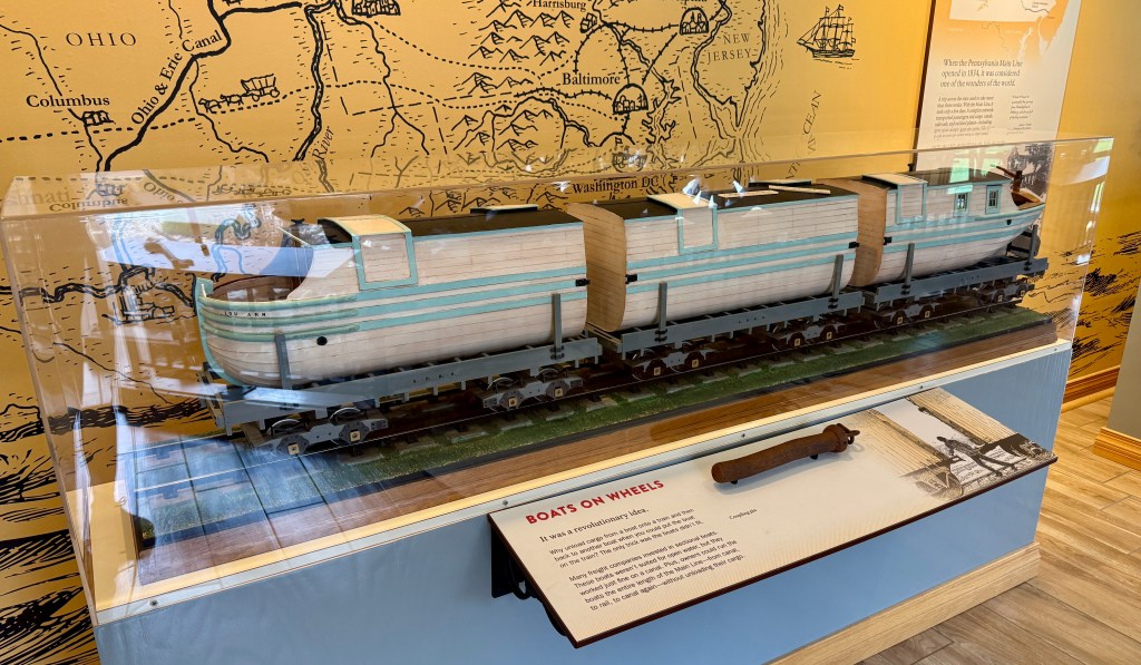 A display case featuring a model of a train designed to resemble a boat, showcasing its unique 'boats on wheels' concept, against a backdrop of a historical map.