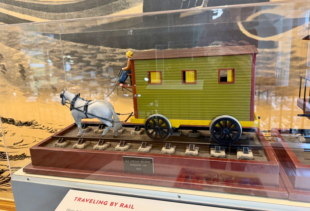A model of an old passenger car from the Old Portage Railroad, featuring a horse-drawn carriage and a seated figure. The car is green with a red roof and has four wheels, displayed on a set of model railroad tracks.