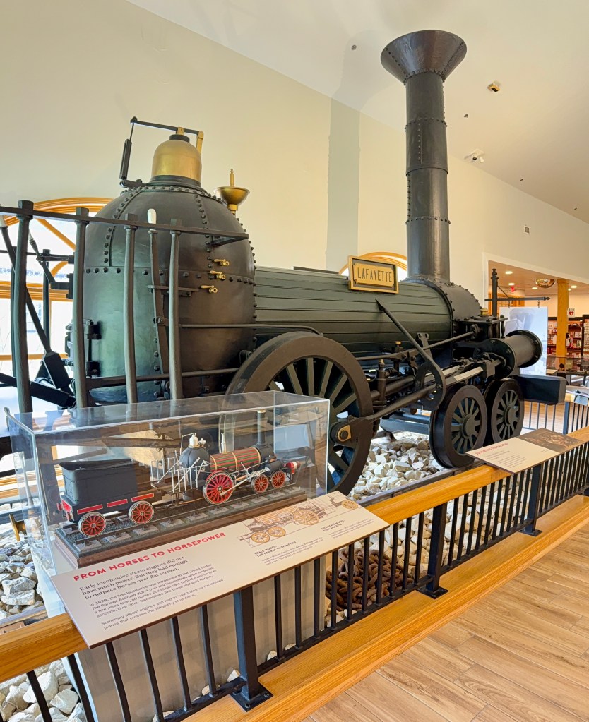 A vintage steam locomotive named 'Lafayette' displayed indoors, with a glass case containing a miniature model of an early train and informational text below.