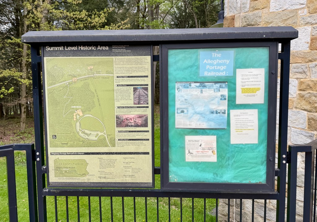 Signboard displaying a map and information about the Summit Level Historic Area and the Allegheny Portage Railroad, surrounded by green trees and grass.