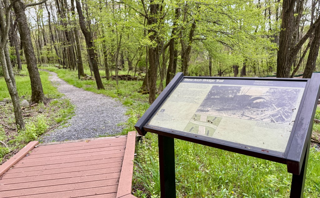 A wooden informational sign for the Skew Arch Bridge Trail in a green forest, with a winding gravel path in the background.