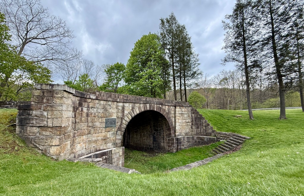 A historic stone arch bridge with steps on the side, surrounded by grassy areas and trees under a cloudy sky.