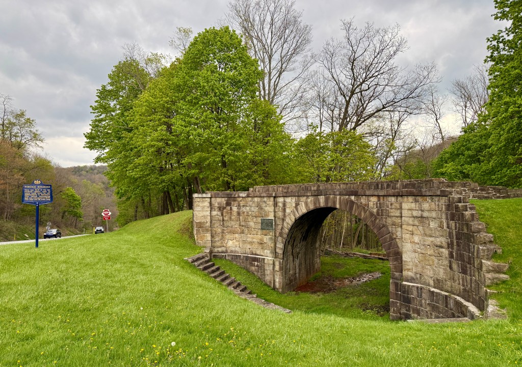 A historic stone arch bridge surrounded by greenery, with a nearby historical marker and a road in the background.