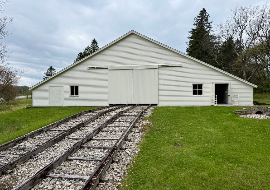 A large white barn with a peaked roof, featuring double doors and adjacent railroad tracks leading to the entrance, set against a cloudy sky and surrounded by greenery.