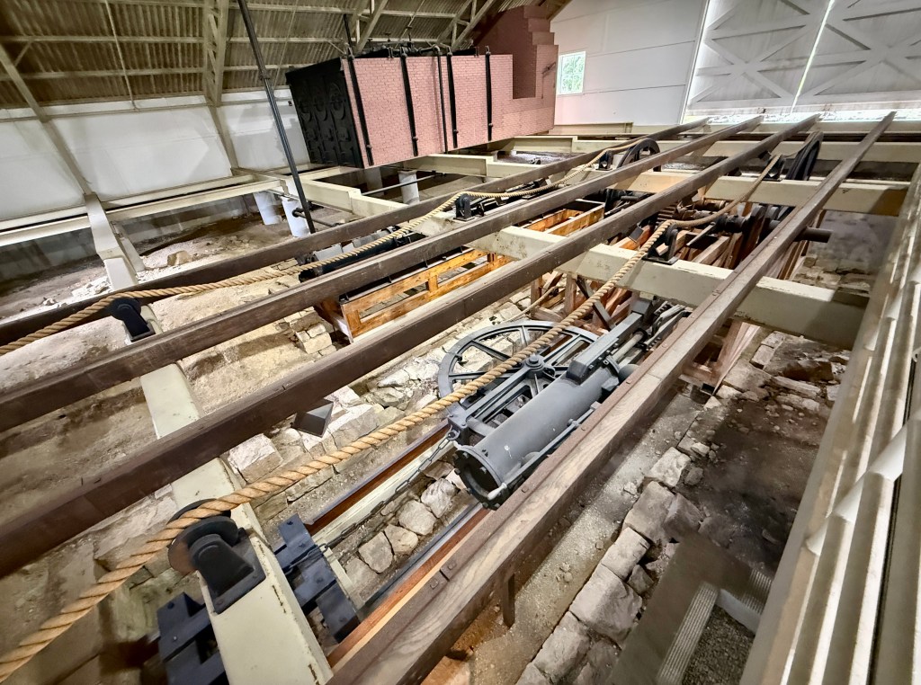 An overhead view of a historical mechanical system featuring wooden beams, ropes, and pulleys, set in a rustic indoor environment.