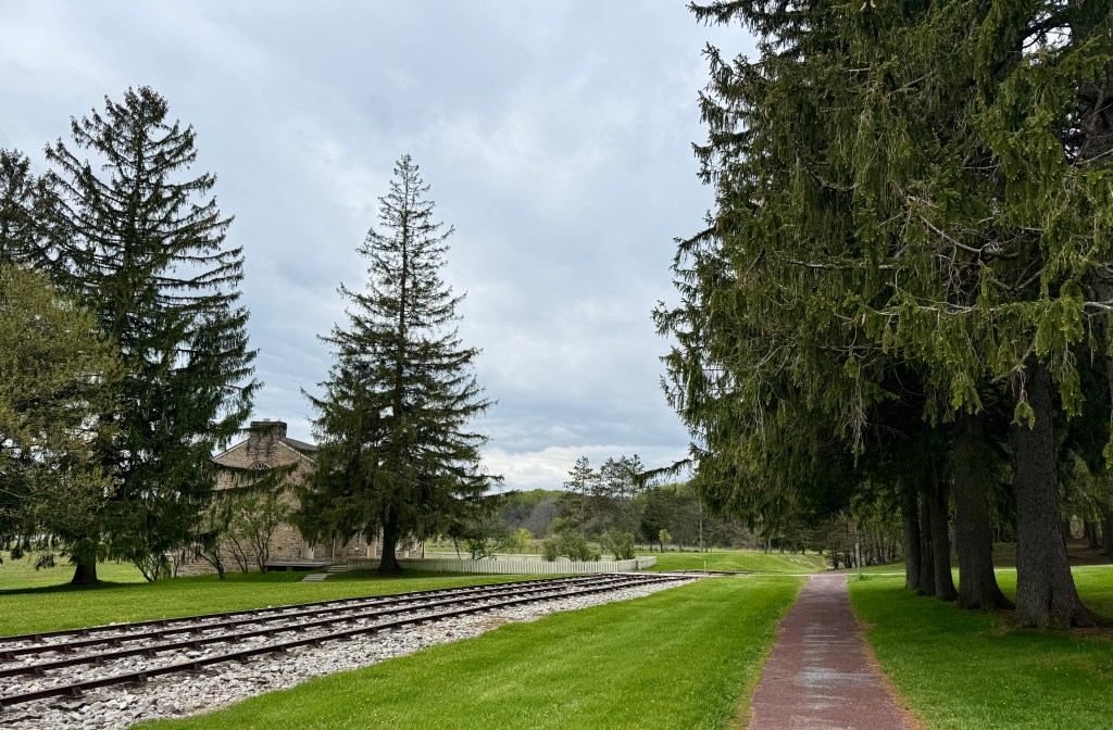 A scenic view of a park featuring lush green grass, tall evergreen trees, and a pathway beside railway tracks. In the background, there is a historic stone building under a cloudy sky.