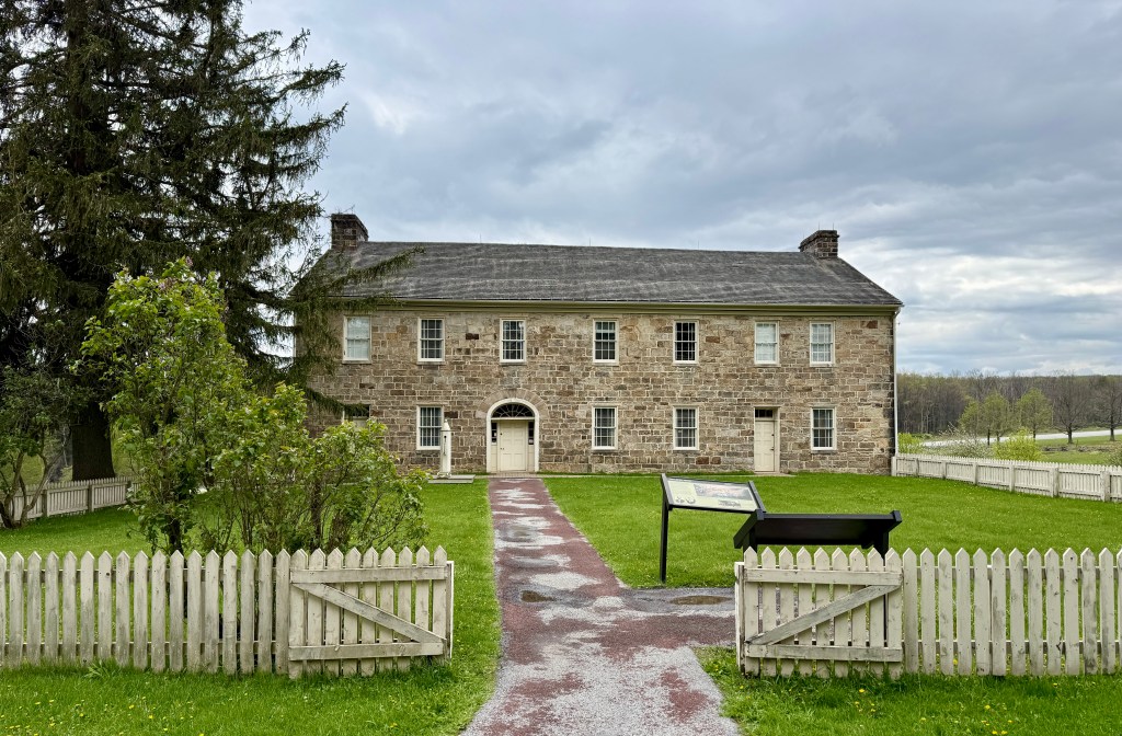 Historic stone building with a grassy lawn and white picket fence, under a cloudy sky.