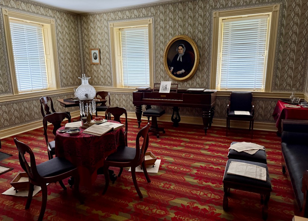 A vintage room featuring patterned wallpaper, a red patterned carpet, a round dining table with a lace tablecloth, and a decorative lamp. A grand piano is present along with two windows providing natural light, and a portrait hanging on the wall.