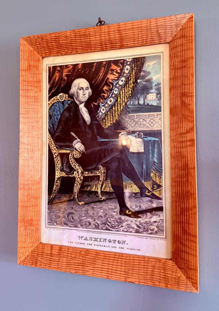 Framed portrait of George Washington seated in an ornate chair, dressed in formal attire, against a backdrop of draped fabric and a scenic view.