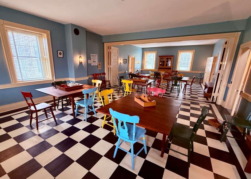 A spacious room with a checkered floor, featuring several wooden tables and colorful chairs in blue, yellow, red, and green. Large windows provide natural light, and there are light blue walls with decorative elements.