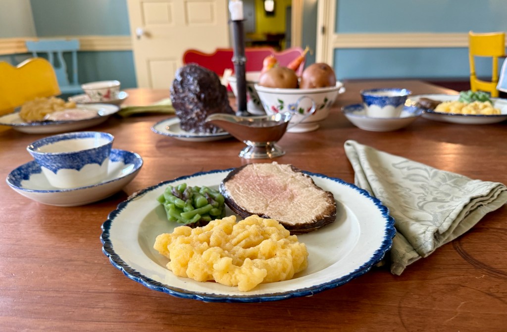 A dining table set with various plates of food, including a portion of macaroni and cheese, green beans, and roast beef. In the background, cups and fruit are visible, all presented on a wooden table with a decorative tablecloth.