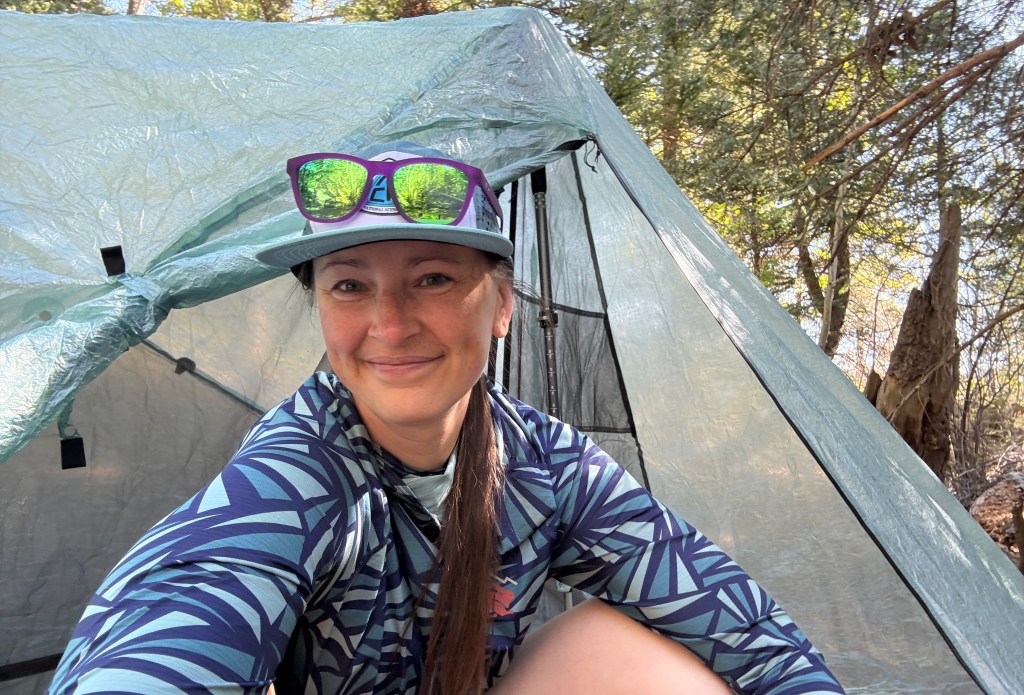A person smiling inside a tent surrounded by trees, wearing sunglasses and a patterned shirt.