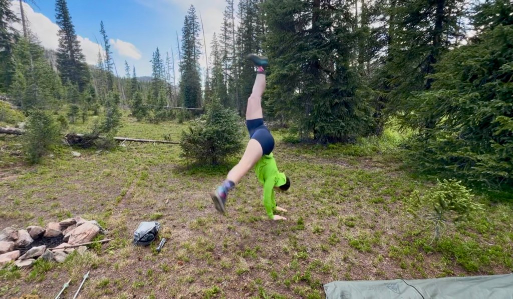 A person wearing a bright green shirt and black shorts performing a handstand in a forested area with trees and a camping setup nearby.