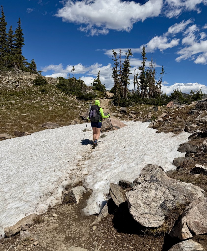 Hiker wearing a bright green jacket walking on a snow-covered trail surrounded by rocks and trees under a blue sky with clouds.