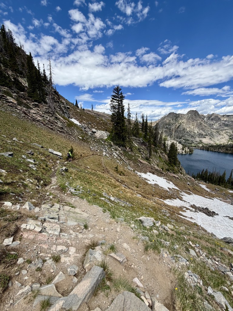 A hiking trail winds through rocky terrain leading down to a scenic lake surrounded by mountains under a blue sky with fluffy clouds.