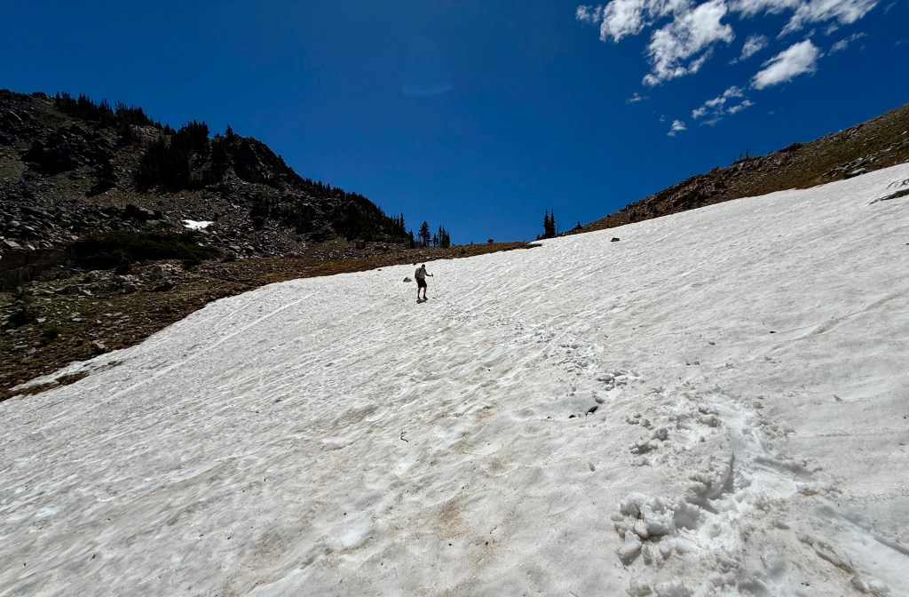 A person hiking up a snowy slope under a clear blue sky, surrounded by rocky terrain and sparse trees.