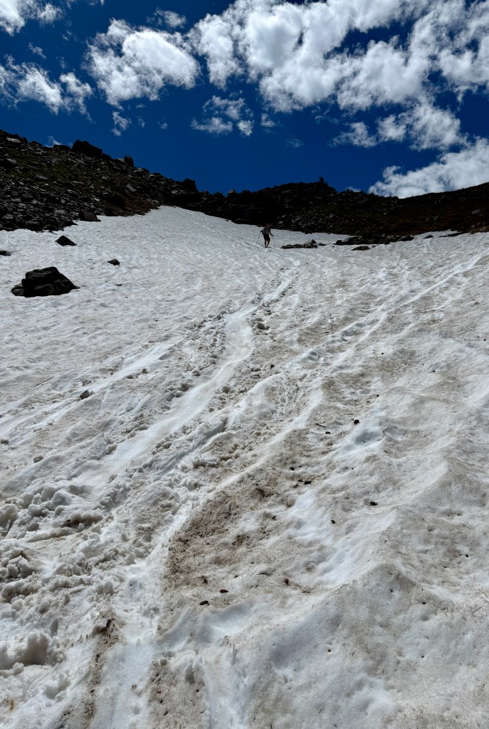 A person walking uphill on a snow-covered slope under a bright blue sky with scattered clouds.