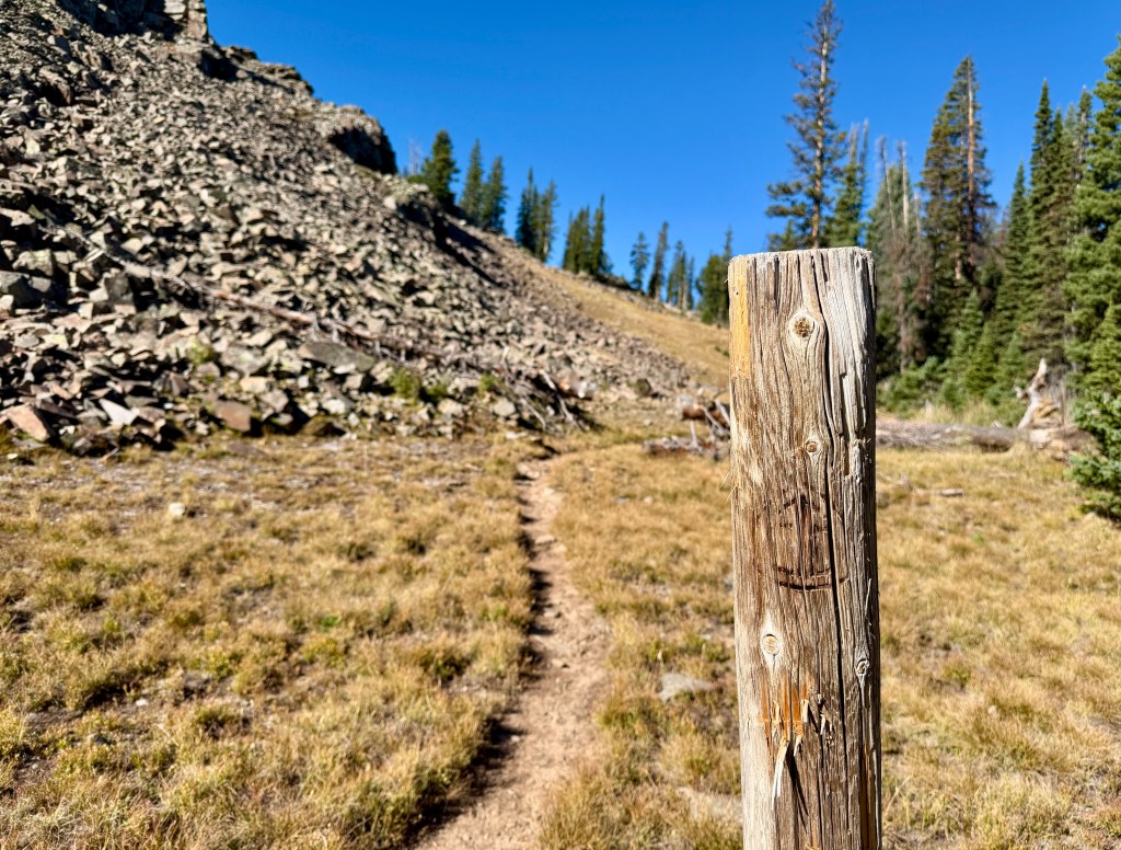 A wooden trail marker stands in the foreground, with a dirt path winding into a grassy area and rocky terrain in the background, under a clear blue sky.