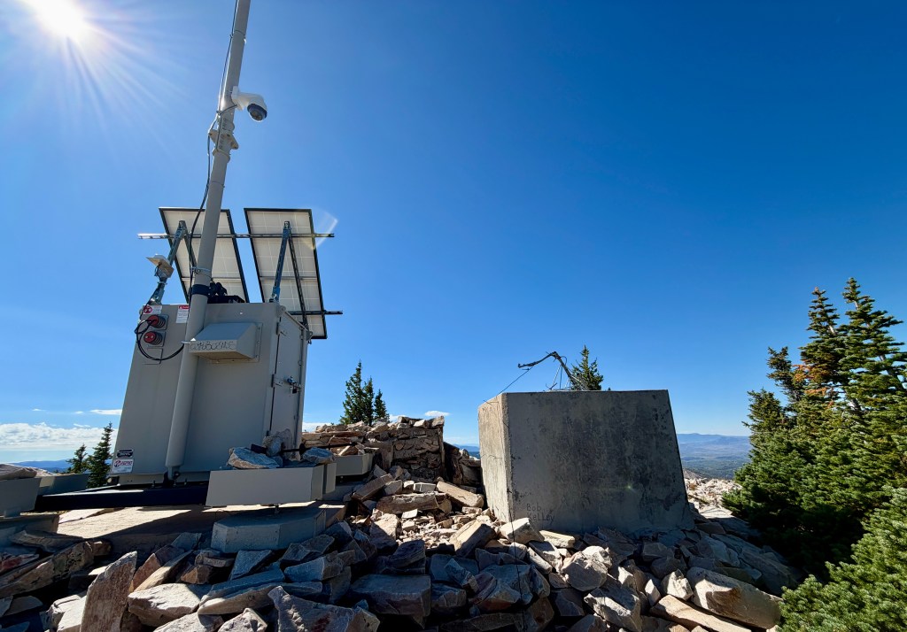 A solar-powered weather monitoring station on a rocky mountaintop, surrounded by pine trees and under a clear blue sky.