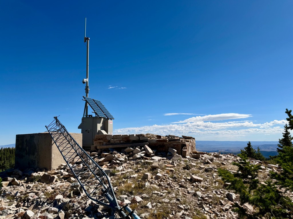 A weather station on a rocky mountaintop with a solar panel, antennas, and scattered boulders under a clear blue sky.