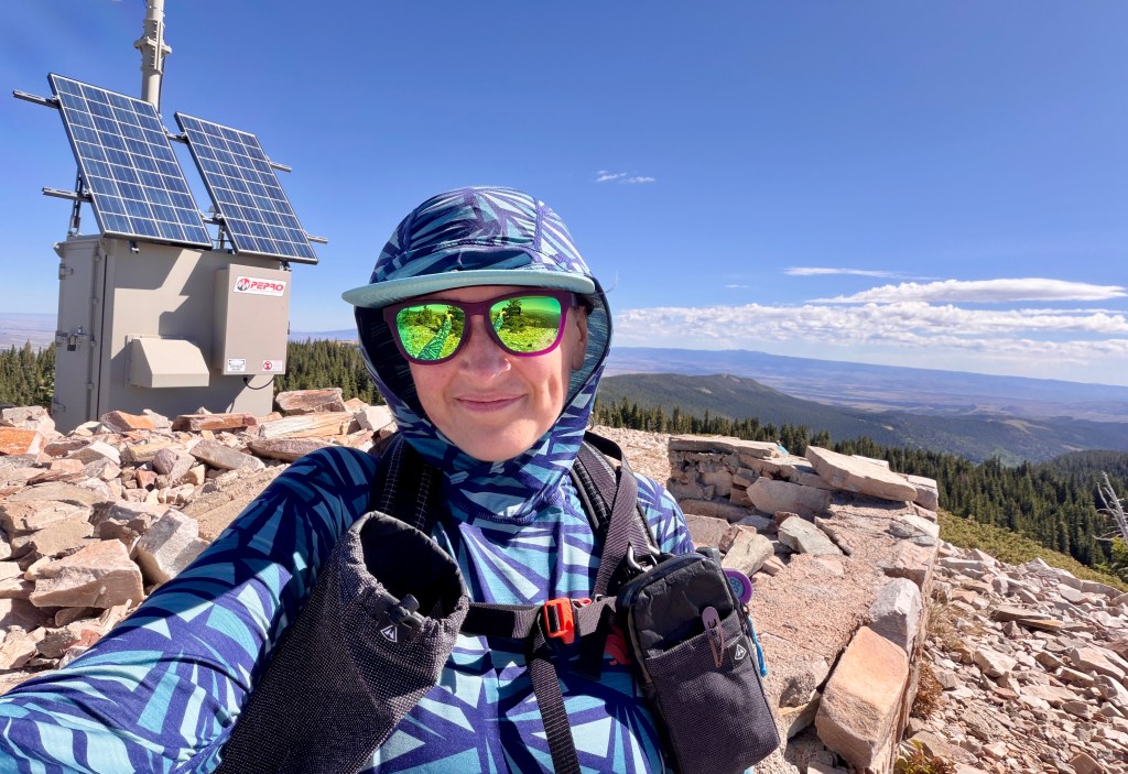 A person wearing a colorful patterned hoodie and sunglasses poses for a selfie on a mountaintop, with solar panels and a scenic view of mountains and trees in the background.