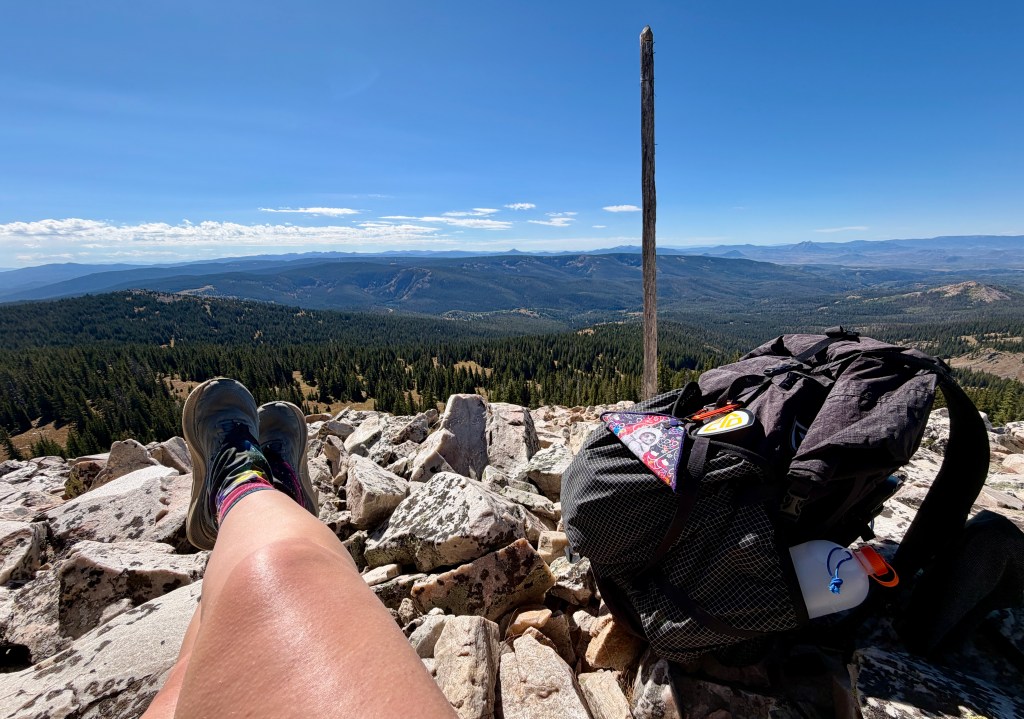 View from a mountaintop showing a person's legs with hiking boots resting on rocks, a backpack nearby, and a mountainous landscape in the background under a blue sky.