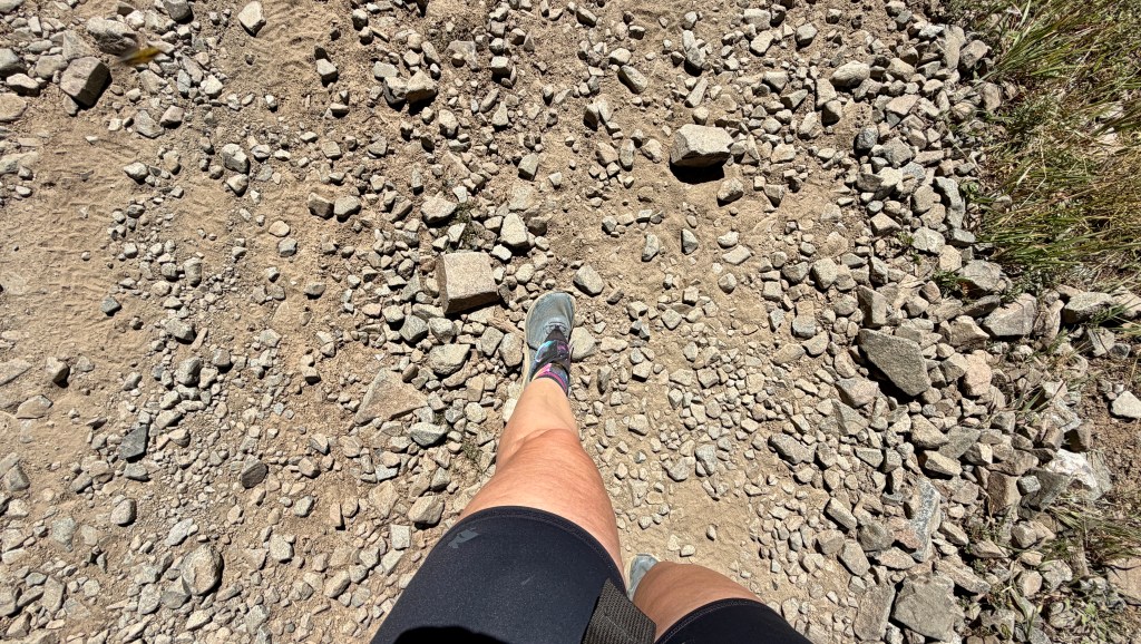 A close-up view of a person's leg and foot wearing a sneaker on a rocky trail, with loose stones and dirt visible on the ground.