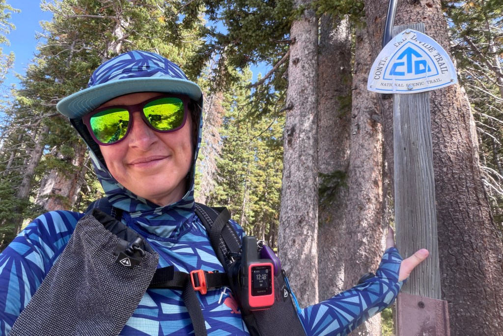 A person wearing a blue patterned outfit and sunglasses stands next to a sign for the Continental Divide Trail, surrounded by tall trees and a clear blue sky.
